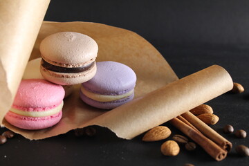Three multi-colored macaroons lie on crafted brown paper next to lie almond sticks cinnamon and coffee on a black background with place for text and with a copyspace
