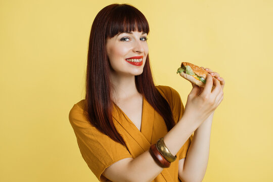 Emotional smiling happy beautiful red haired pretty woman in yellow blouse on yellow studio background, holding fresh tasty cheese burger. Fast food, jank food concept.