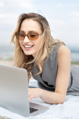 Young stylish woman is working remotely on the laptop computer lying on the beach with a big smile, talking on zoom video chat call 