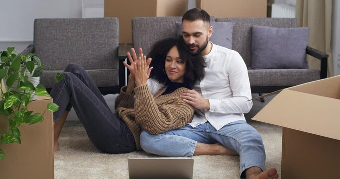 Tired young people african american curly woman and white caucasian man sit hugging on floor near cardboard boxes and houseplants rest after moving to new house apartment flat, buying real estate