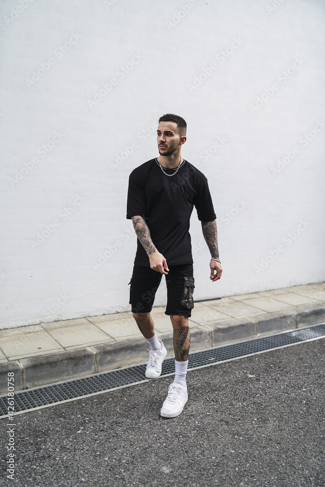 Poster Young Caucasian male from Spain with tattoos wearing a black shirt and shorts posing on a street