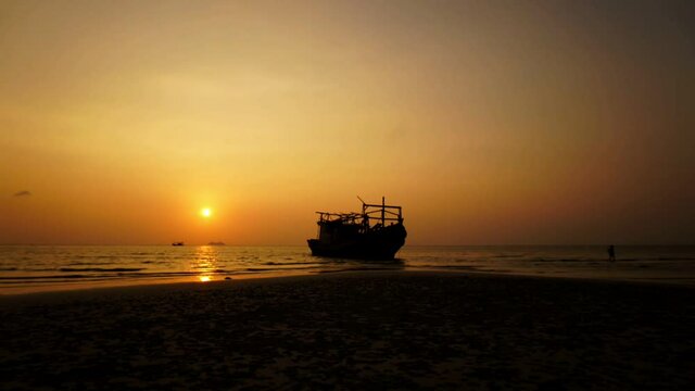 Sun goes down while people walk over the beach in Thailand. Timelapse of the sunset behind the standing boat at the seashore. Theme of vacation and travelling near the ocean.