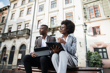 Low angle view of two afro american business colleagues sitting on wooden bench on city street and...