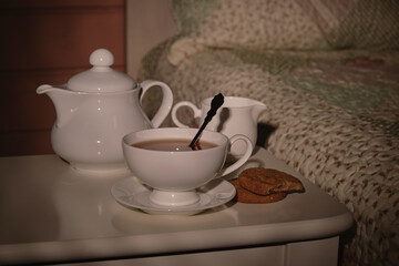 Fototapeta na wymiar A cup of tea, a kettle and a plate of cookies on a small table by the bed in a small cozy bedroom in the morning sunlight. Everything for a good mood. High quality photo