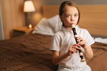 Close-up portrait of talented adorable little girl playing flute sitting on bed in bedroom, looking at camera. Child practices playing musical instrument at home.