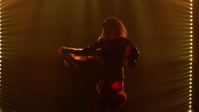 A passionate woman in a gypsy costume whirls dancing and waving her long skirt. A dancer is silhouetted in a gypsy folk dance in a dark smoky studio with yellow light. Close up.