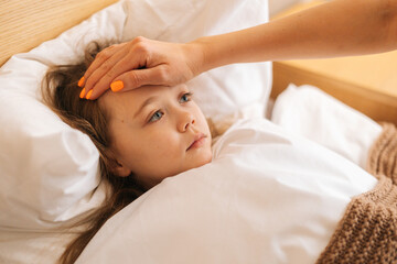 Close-up view of young mother measures temperature touches forehead of sick daughter lying in bed at home. Parent caring for sick child lying in bed with flu. Concept of children health.