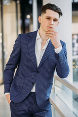 portrait of sexy young businessman in navy blue suit holding chin, looking at camera and posing, holding hand in pockets