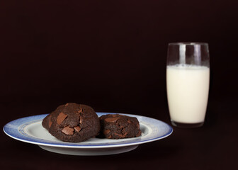 Plate on dark brown background with three chocolate cookies with a glass of milk aside