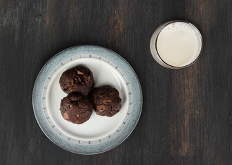 Plate on wooden table with three chocolate cookies with a glass of milk aside; top view.