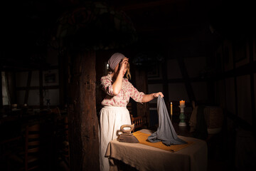 Fototapeta na wymiar Eine Frau Mitte Vierzig, ende Achtzehn Jahrhundert bügelt mit dem alten verrosteten Bügelleiser die Bekleidung. Sie ist überarbeitet. Die Frau hat Kopftuch und Schurze an. Die Gebäude ist altmodisch 