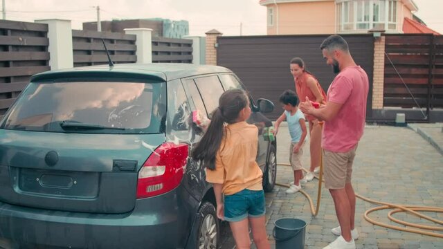 Full shot of elementary age brother and sister helping their young adult parents to wash car outdoors in backyard on hot summer day