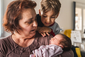 Small caucasian baby newborn infant girl yawing in hands of her grandmother while her older brother...