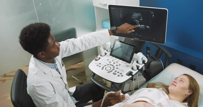 Ultrasound concept. Top angle view of young Caucasian pregnant woman lying on the couch, while high skilled male African American doctor making ultrasound scan and showing the baby on the screen