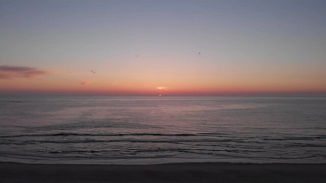 DRONE AERIAL FOOTAGE: Sea, beach boulders and waves at sunset. Beautiful sunset with flying birds. Waves crashing on sandy beach. Beach with sand dunes and dusk sky in Ofir, Fao, Portugal. 