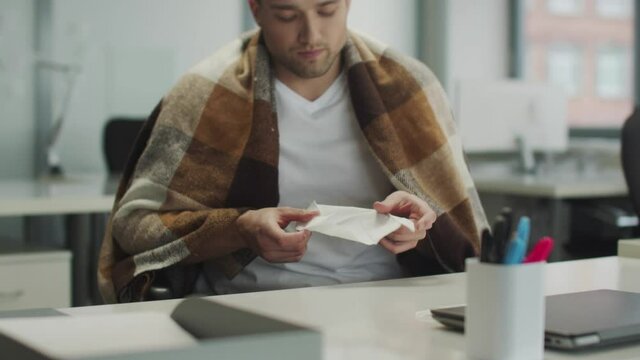 A man with a blanket on his shoulders is sick at home or in the office. He blows his nose into a disposable paper handkerchief, slowmotion.