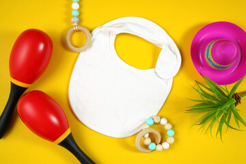 Cinco de Mayo baby bib flatlay on a festive yellow table background, styled with colorful maracas and sombrero hats. White product mock up with negative copy space for your text or design here.
