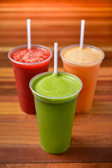 Fruit juices in red, orange and green colors in plastic cups and straws on a dark wooden table.