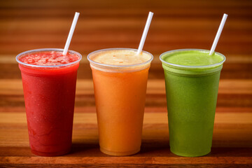 Fruit juices in red, orange and green colors in plastic cups and straws on a dark wooden table.