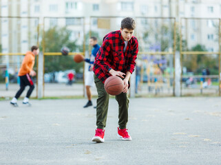 Cute young boy plays basketball on street playground. Teenager in red  flannel checked shirt with orange basketball ball outside. Hobby, active lifestyle, sport activity for kids.