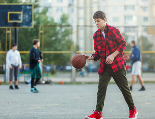 Cute young boy plays basketball on street playground. Teenager in red flannel checked shirt with orange basketball ball outside. Hobby, active lifestyle, sport activity for kids.