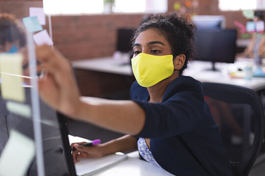 Mixed rase businesswoman wearing mask sticking note on glass wall