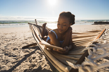 Happy african american woman lying in hammock on beach using tablet