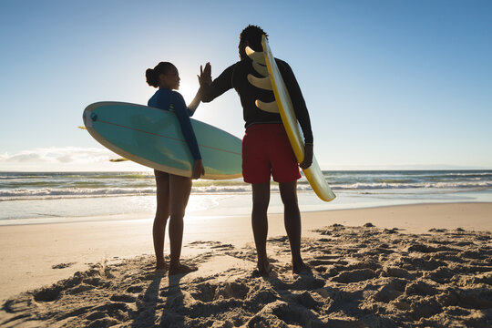 Happy african american couple on the beach carrying surfboards high five