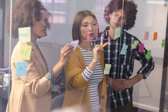 Diverse group of creative colleagues brainstorming writing notes on glass wall in meeting room