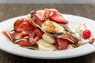 Mouthwatering plated of crepes piled with sliced bananas and strawberrries topped with whipped cream and syrup.