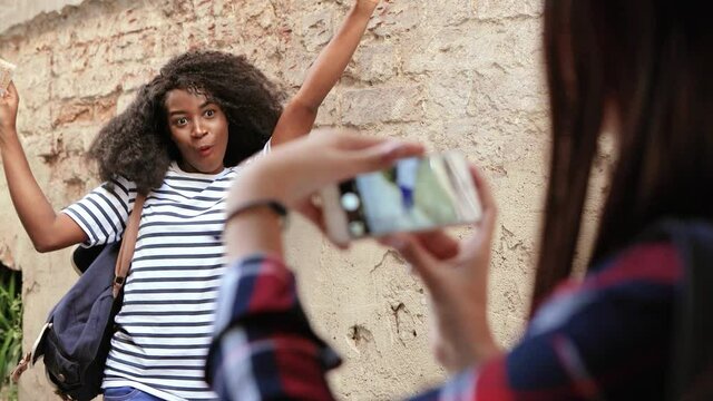 Young beautiful african american girl tourist with curly hair and backpack posing for photos while her friend is taking pictures on smartphone.