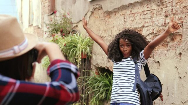 Young beautiful african american girl with curly hair and backpack posing for photo raising her arms up standing on background of old stone wall.