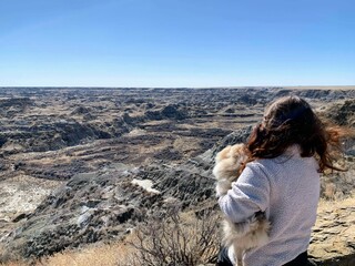 Fototapeta na wymiar A woman holding her small dog admiring a beautiful panoramic view of the badlands in Dinosaur provincial park, Alberta, Canada. Full of valleys of hoodoos and coulees.