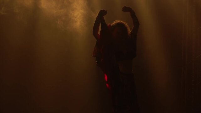 A passionate woman in a gypsy costume whirls dancing and waving her long skirt. A dancer is silhouetted in a gypsy folk dance in a dark smoky studio with yellow light. Close up. Slow motion.