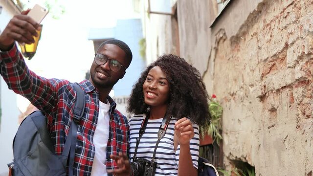 Young African American couple of tourists walking around the city and taking photos for social networks. Handsome guy with backpack holding smartphone, girl sending air kisses.