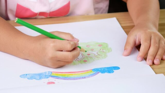 Close up hand of child drawing a rainbow and tree with colored pencils at the table at home.