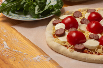 Raw pizza on baker paper on the wooden table. Preparing homemade uncooked cheese pizza with tomato sauce, tomato cherry, sausage and dough. 