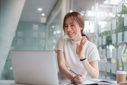 Happy Asian young business woman looking at computer screen, waving hello. using meeting online app, work from home, working remotely, social distancing, new normal concept.