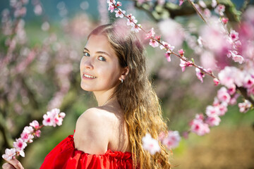 Close up spring portrait of young beautiful smiling lady wearing red dress and posing in park with blooming trees