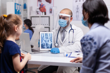 Professional therapeutist showing radiography on tablet pc during coronavirus pandemic. Pediatrician doctor in protection gloves and mask providing healtcare services in medical clinic