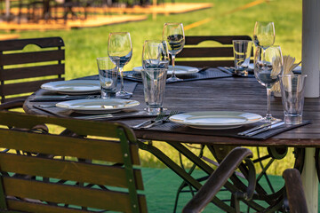 Banquet service, outdoor catering in the park. Table setting. White plates, glasses for wine and drinks, forks, knives. Empty wine glasses on a wooden table in an outdoor cafe.