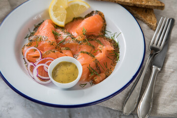Gravlax Graved Lachs, ein roh gebeizter Fisch mit Dill auf Emaille Teller mit Hovmästarsås Senf Soße, Toast, Zwiebel Ringe, Zitrone und Leinen Tuch auf hell Marmor Hintergrund