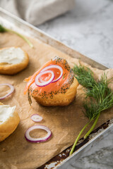 Gravlax Graved Lachs, ein roh gebeizter Fisch mit Dill auf Brötchen mit Frischkäse,  Zwiebelringe auf Backblech mit Backpapier und hell Marmor Hintergrund
