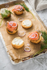 Gravlax Graved Lachs, ein roh gebeizter Fisch mit Dill auf Brötchen mit Frischkäse,  Zwiebelringe auf Backblech mit Backpapier und hell Marmor Hintergrund