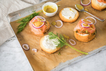 Gravlax Graved Lachs, ein roh gebeizter Fisch mit Dill auf Brötchen mit Frischkäse, Hovmästarsås Senf Soße, Zwiebelringe, auf Backblech mit Backpapier und hell Marmor Hintergrund