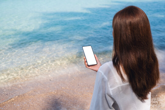 Rear view mockup image of a woman holding mobile phone with blank desktop screen while sitting on the beach