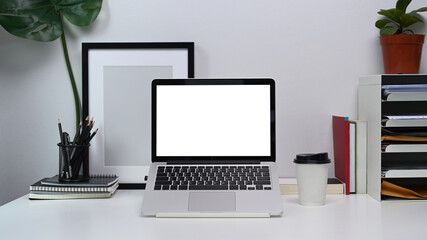 Mock up computer laptop with blank screen, notebook, empty photo frame and coffee cup on white office desk.