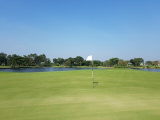 A golf course with a white flag on its hole and a pond and clear sky.