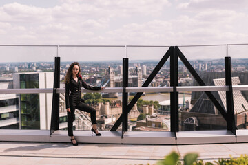 Stylish business lady in a classic black suit. In full growth. Beauty. Background buildings, city landscape. Sky terrace. Skyscrapers.