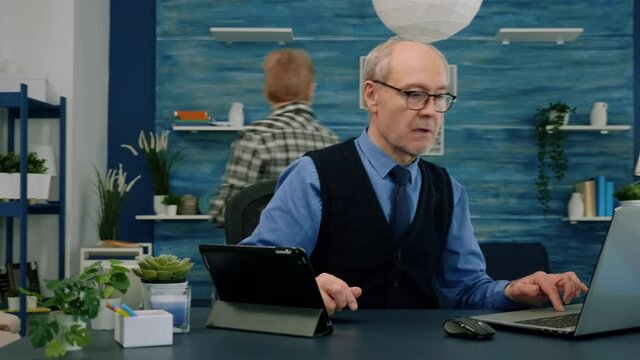 Elderly man using tablet and laptop in same time compering financial graphs working from home sitting in workplace. Busy focused old employee writing, searching, analysing reports
